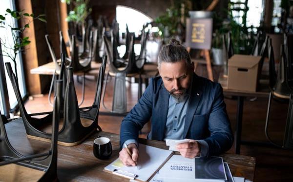 A man with gray hair in a bun, wearing a blue suit, sits doing paperwork at a table surrounded by other tables with the chairs placed on top of them.