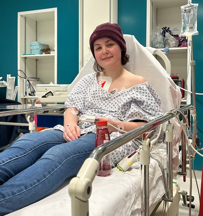 Imali Chislett sitting upright in a hospital bed, wearing jeans and a hospital gown, with lots of tubes attached to her